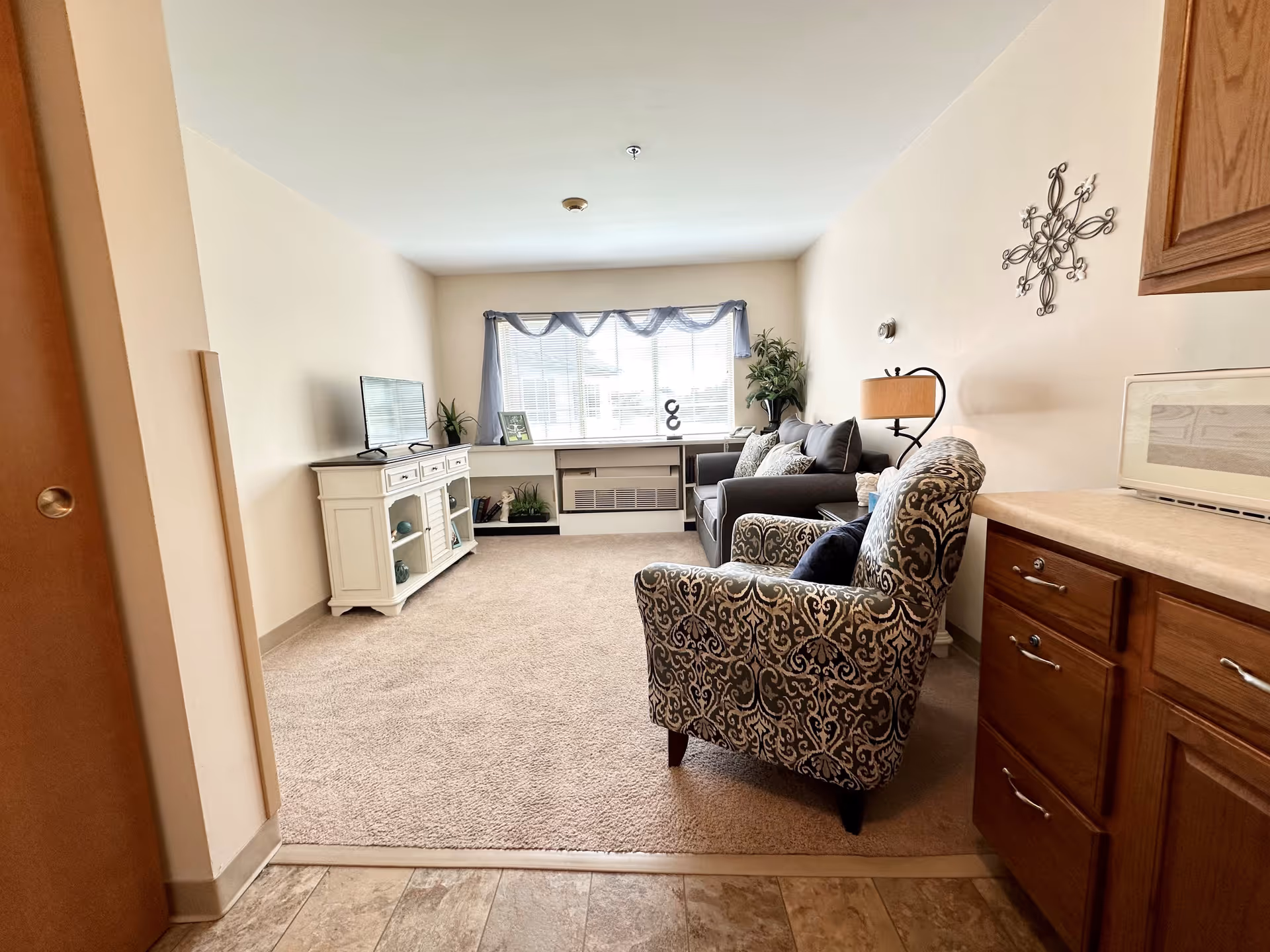 A cozy living room in a senior living community featuring a patterned armchair, a dark gray sofa with cushions, a white TV stand with a flat-screen TV, decorative plants, a window with sheer curtains, and wooden kitchen cabinets on the right side.