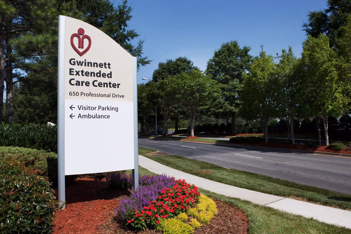 Outdoor view of a sign for Gwinnett Extended Care Center located at 650 Professional Drive, with directions for visitor parking and ambulance. The sign is surrounded by landscaped flowers and bushes, with a road and trees in the background under a clear blue sky.