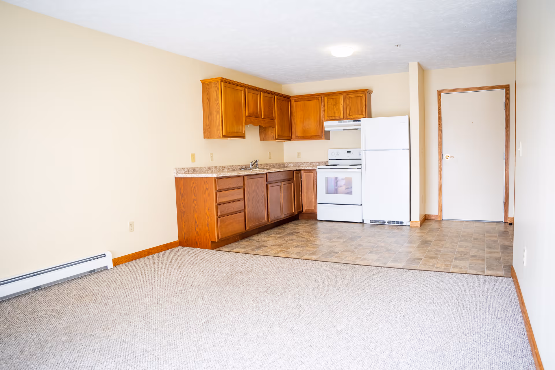 An empty small kitchen area with wooden cabinets, a white stove, and a white refrigerator. The kitchen floor is tiled, and the adjacent living area is carpeted. The walls are painted light beige, and there is a white door with wooden trim in the background.