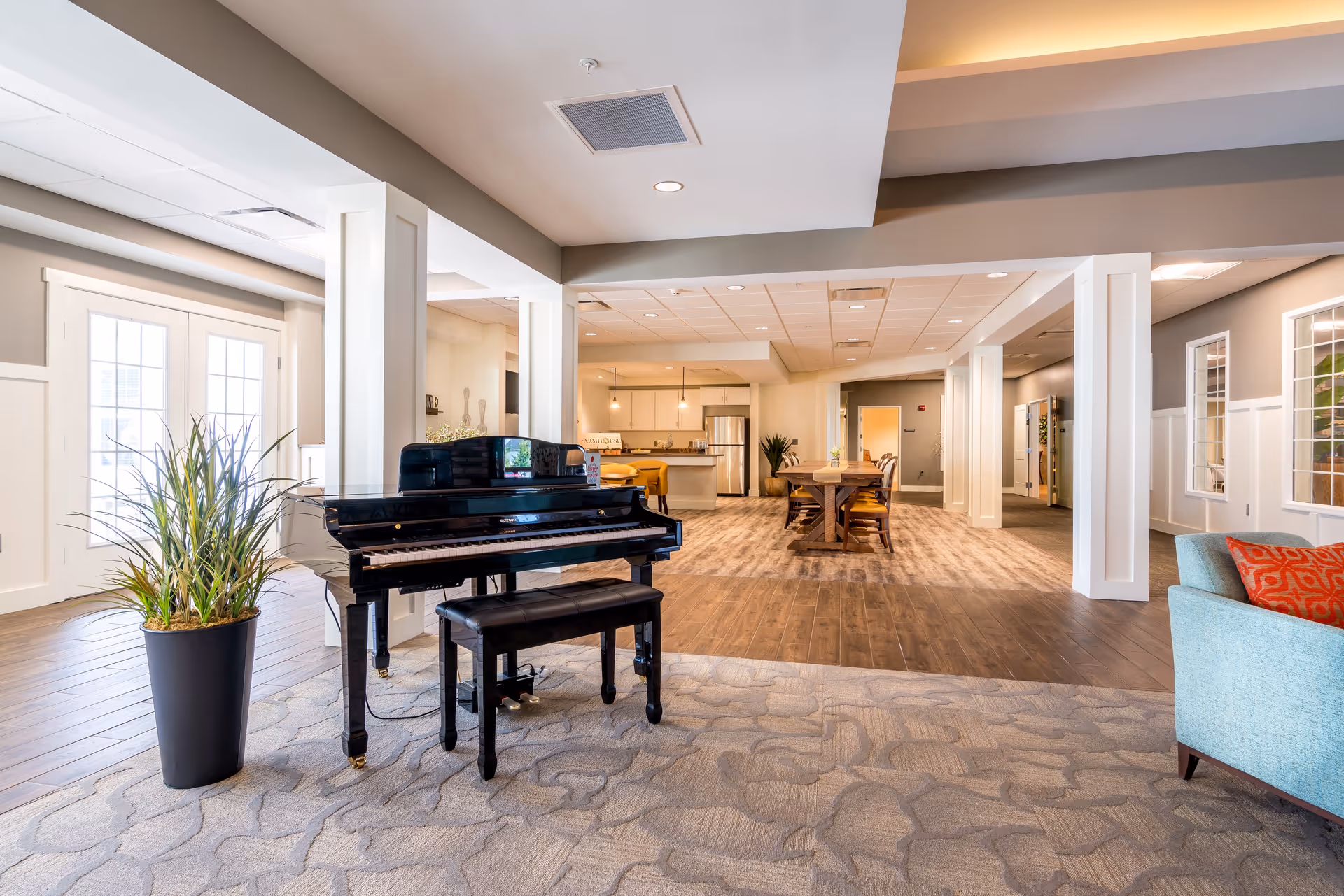 A spacious and well-lit common area in a senior living facility featuring a black grand piano with a matching bench on a patterned carpet. To the left, there is a tall potted plant near large glass doors. In the background, there is a dining table with chairs and a kitchen area with modern appliances. The room has light-colored walls, wooden flooring, and recessed lighting.