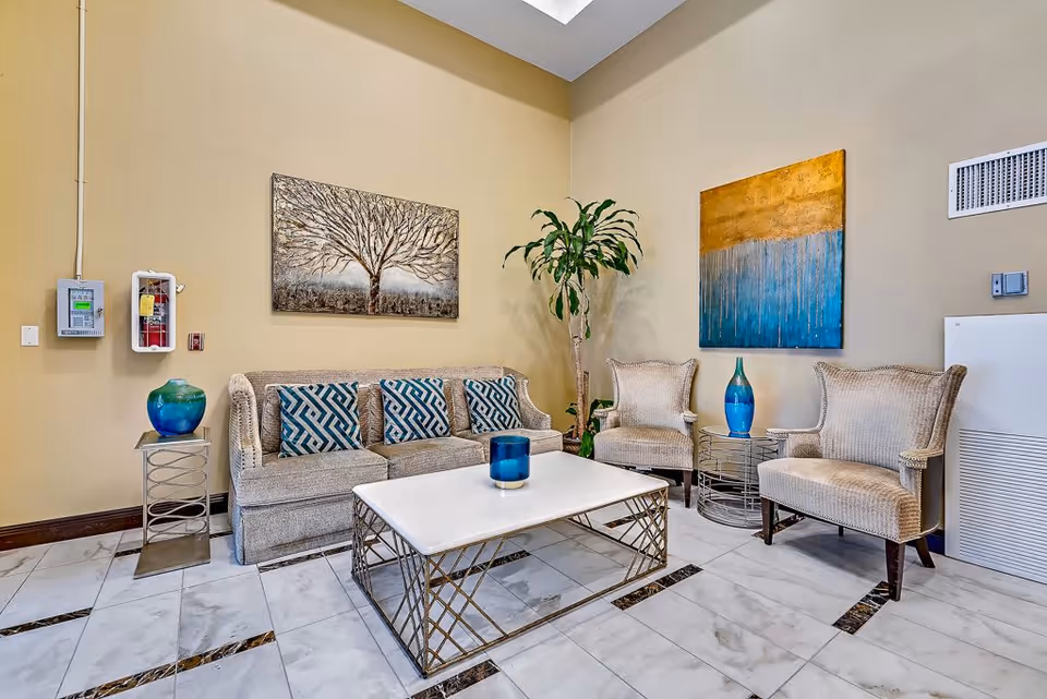 A cozy seating area in a senior living facility with a beige sofa adorned with blue patterned cushions, two beige armchairs, a white marble coffee table with a blue candle, and side tables with decorative blue vases. The walls are painted light beige and decorated with two large paintings, one depicting a tree and the other an abstract design. A tall green plant is placed in the corner, and the floor is tiled with white and dark marble accents.