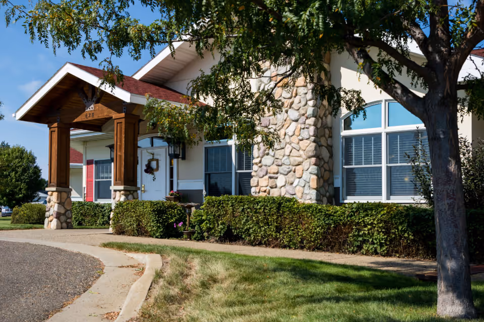 Front entrance of a residential building with stone columns, a wooden portico, large windows, and landscaped shrubs.