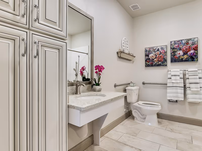 A clean and modern bathroom with a white toilet, a wall-mounted sink with a marble countertop, a large mirror, and white cabinets. The bathroom is decorated with two colorful floral paintings, a small shelf with decorative items, and two striped towels hanging on a towel rack. There is a pink orchid plant on the countertop.