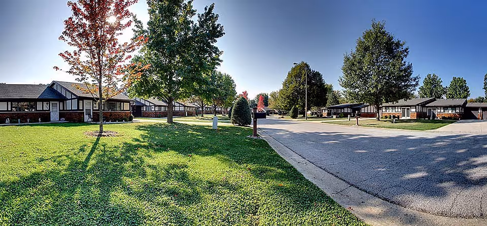 A sunny suburban street view of Villa Senior Care Community showing single-story residential buildings with brick and siding exteriors, green lawns, and several trees casting shadows on the grass and pavement.