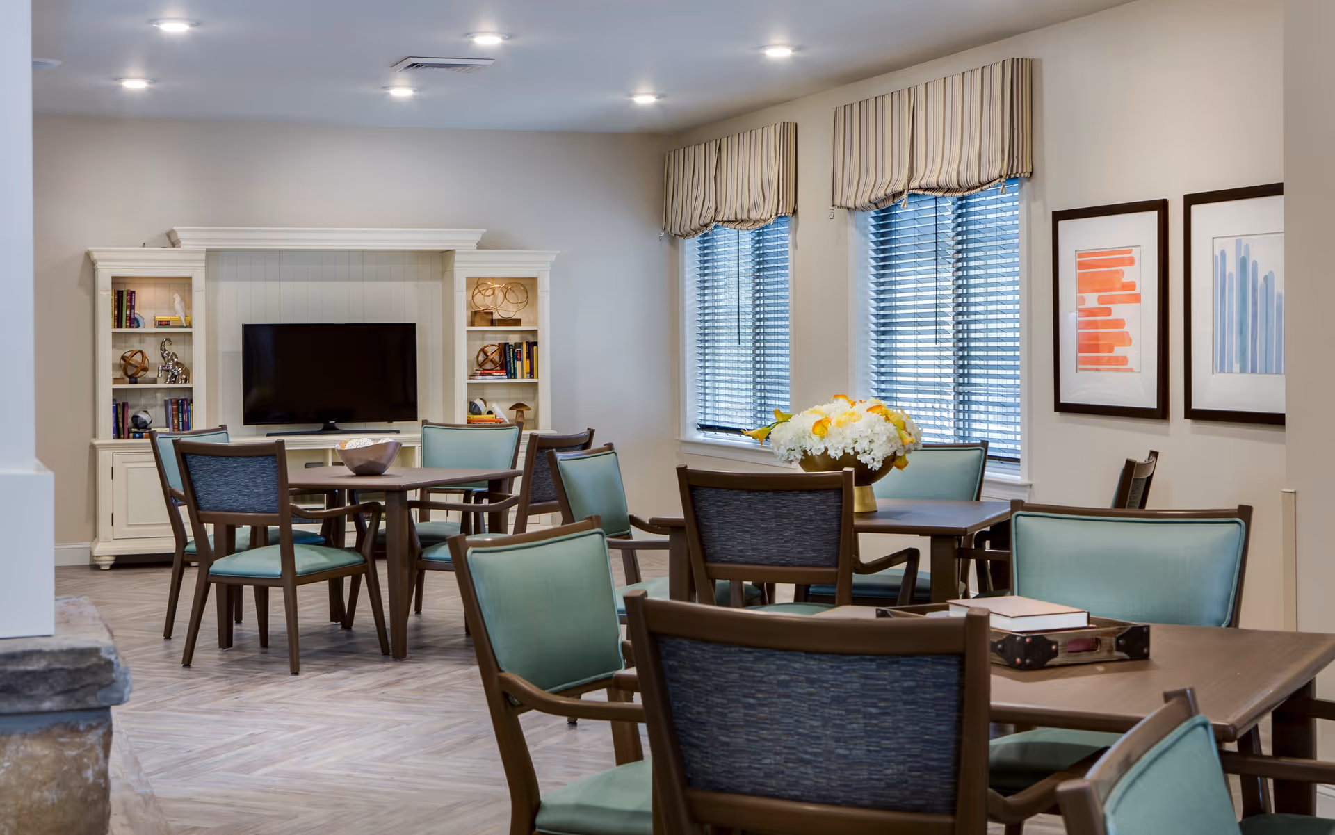A well-lit common area with multiple wooden tables and chairs featuring light blue cushions. There is a white entertainment center with a flat-screen TV and decorative items on shelves. Two windows with striped valances and blinds let in natural light. Two framed abstract artworks hang on the wall.