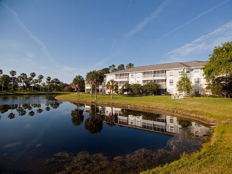 White three-story senior living building beside a pond with palm trees and its reflection under a clear blue sky.