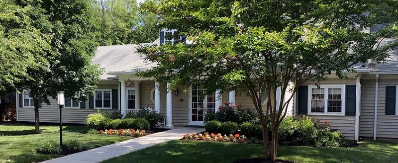 Front exterior view of a single-story residential building with beige siding, white trim, and a gray roof. The entrance features white columns and a glass door. There is a well-maintained lawn with a concrete walkway, flower beds, and several trees and shrubs in front of the building.
