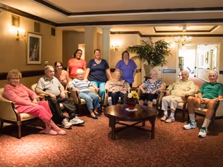 A group of elderly people sitting in a living room area of a senior living facility, arranged in a semi-circle on chairs and sofas around a wooden coffee table with a flower arrangement. The room has warm lighting, carpeted floors, and framed artwork on the walls.