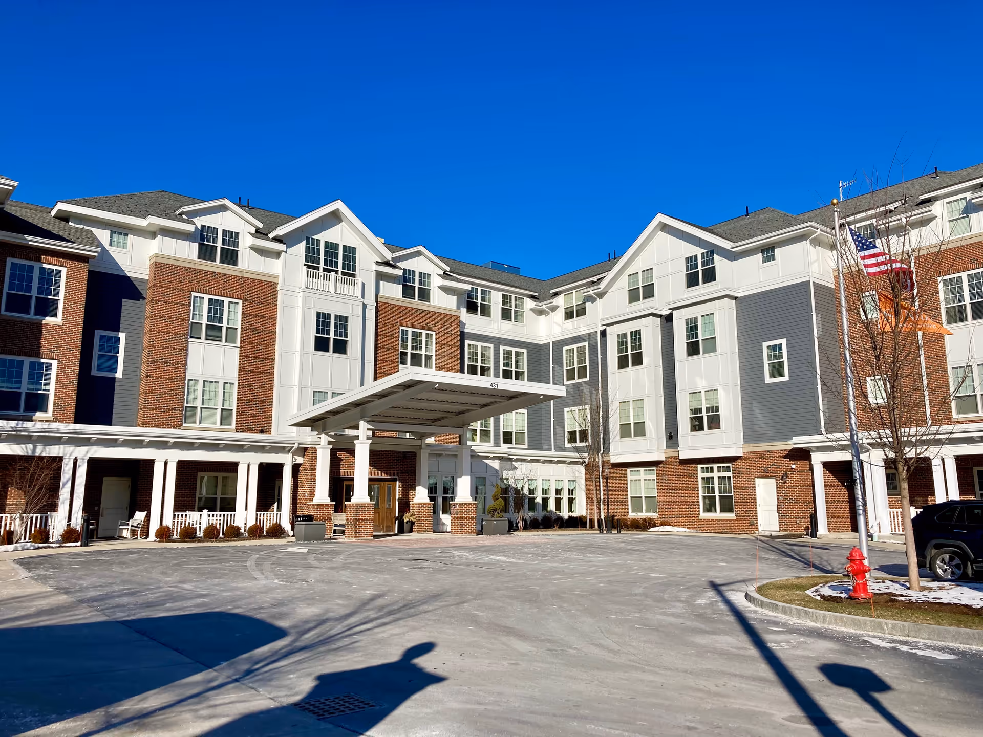 Exterior front view of a multi-story senior living facility building with a covered entrance, brick and white siding facade, multiple windows, an American flag, and a clear blue sky.