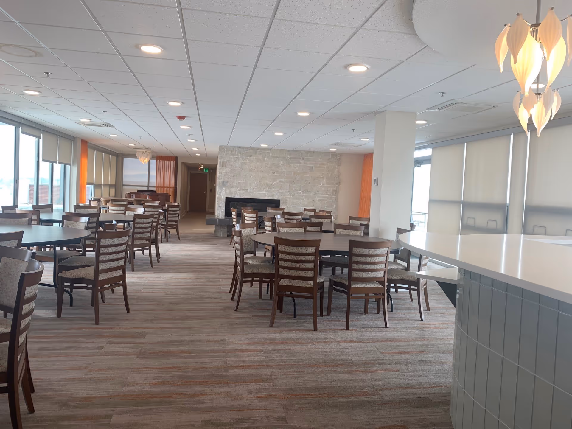 A spacious dining room in an assisted living center with multiple round and rectangular tables surrounded by wooden chairs. The room features large windows with blinds, a stone fireplace on the far wall, and modern ceiling lights. A white countertop with a decorative hanging light fixture is visible on the right side.
