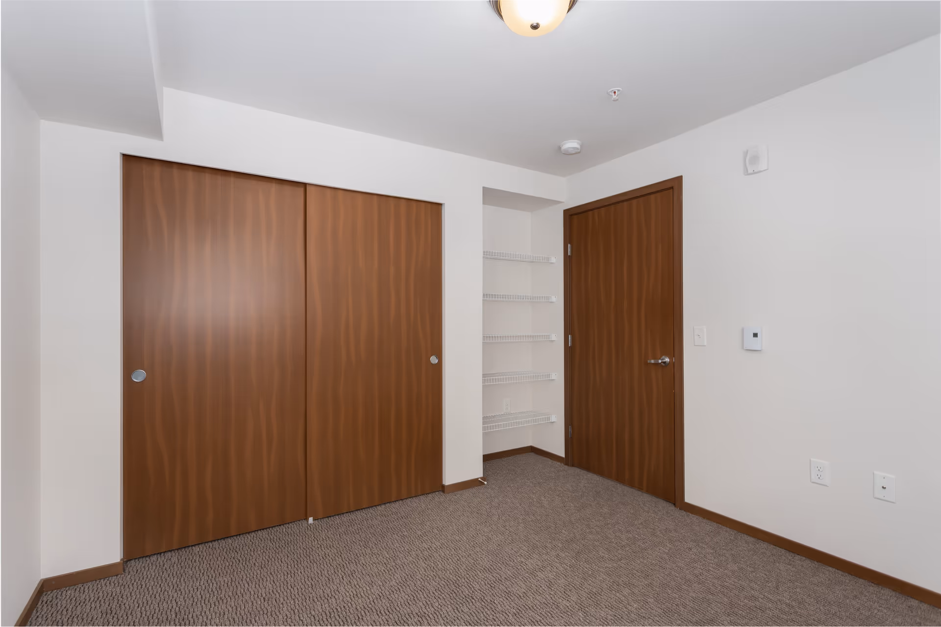 Empty room corner with beige carpet, white walls, a wooden door on the right, and a wooden sliding closet door on the left. There are four white wire shelves in a recessed wall space between the doors.