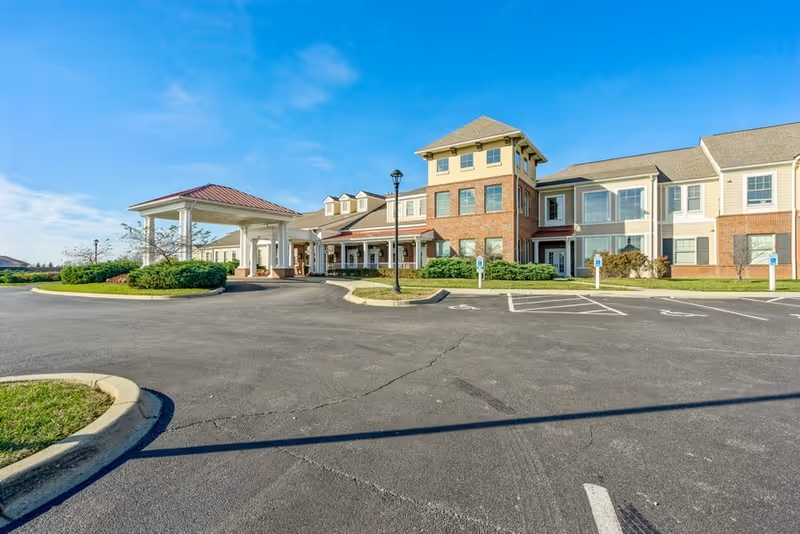 Exterior view of The Enclave of East Louisville senior living facility showing a large two-story building with a covered entrance, multiple windows, and surrounding landscaping under a clear blue sky.