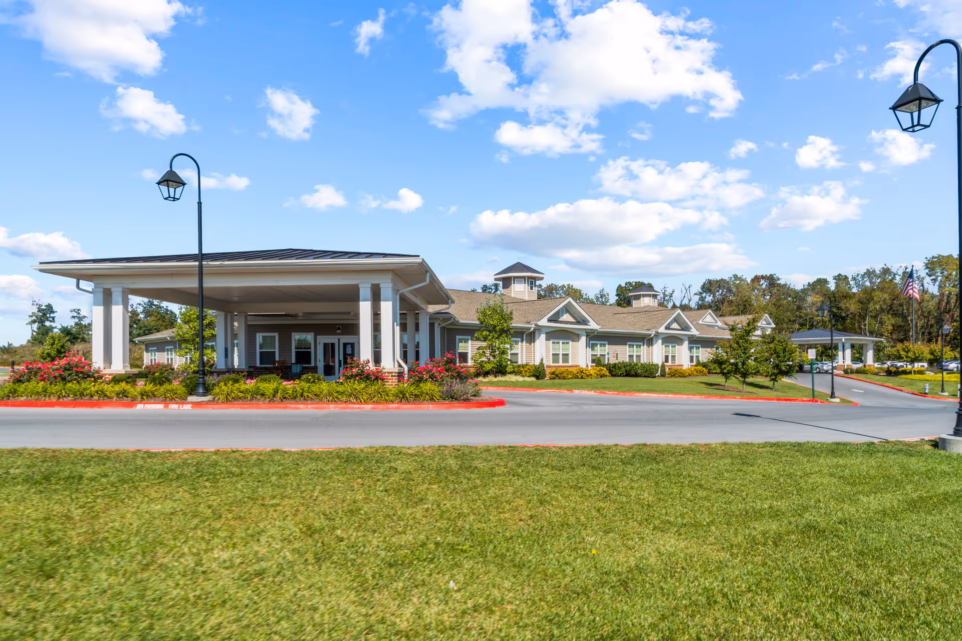 Exterior view of Bellaire at Stone Port Assisted Living & Memory Care facility showing a single-story building with a covered entrance, surrounded by well-maintained landscaping, green grass, and a clear blue sky with scattered clouds.
