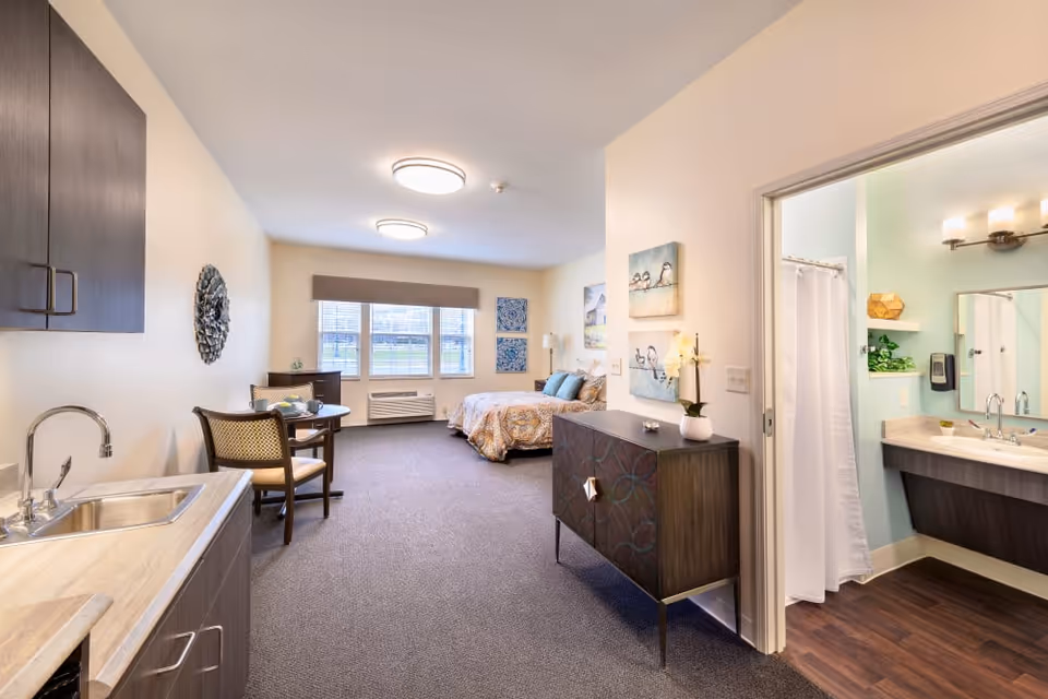 Well-lit studio apartment interior with a bed, kitchenette, small dining table, dresser, and an open bathroom.