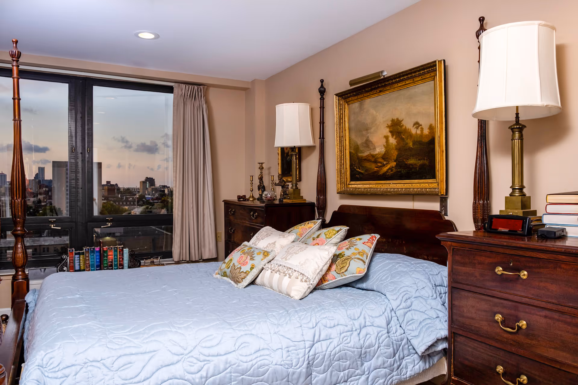A cozy bedroom with a large wooden four-poster bed covered with a light blue quilt and several decorative pillows. There are two wooden dressers on either side of the bed, each with a lamp. A framed painting hangs above the bed. A large window with beige curtains shows a cityscape view at dusk.
