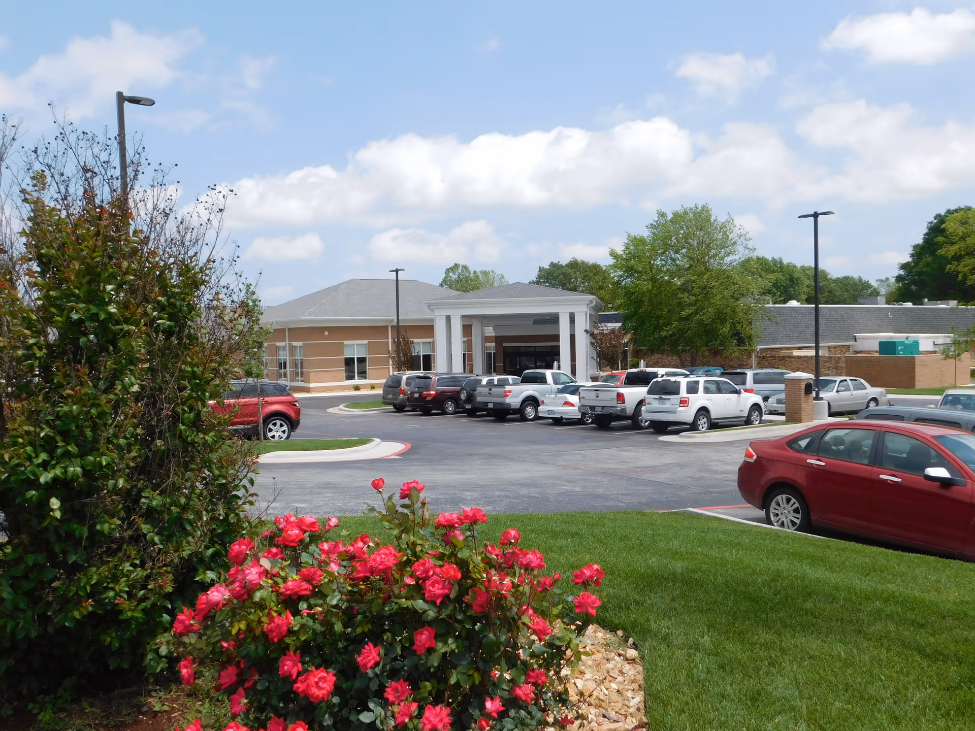 Exterior view of Springfield Rehabilitation & HealthCare Center showing the front entrance with a covered drop-off area, a parking lot with several cars, green grass, blooming red flowers, and trees under a partly cloudy sky.