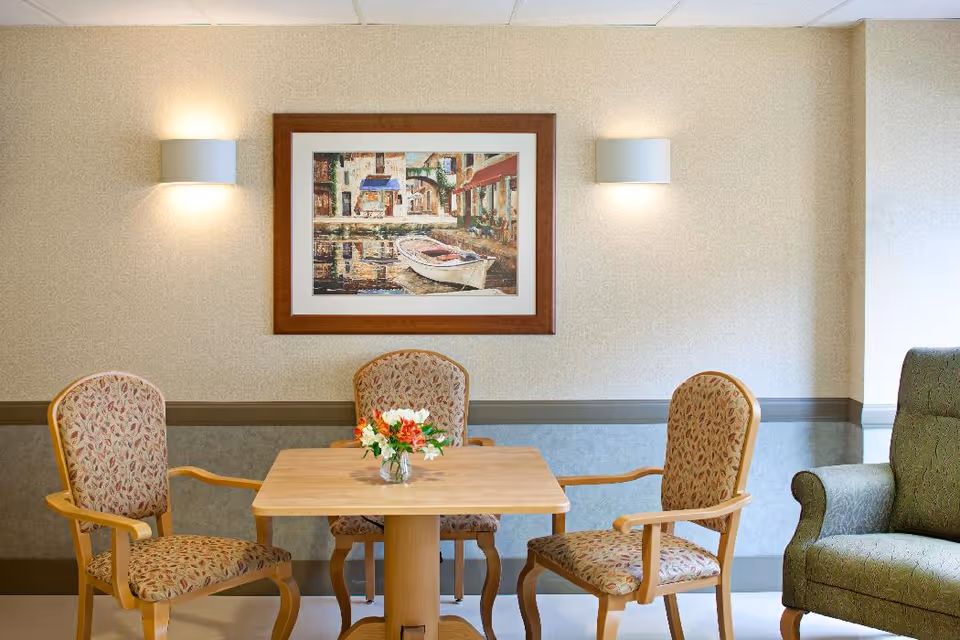 Small dining area with a wooden table, three upholstered chairs, a vase of flowers, framed artwork and wall sconces on a neutral wall.