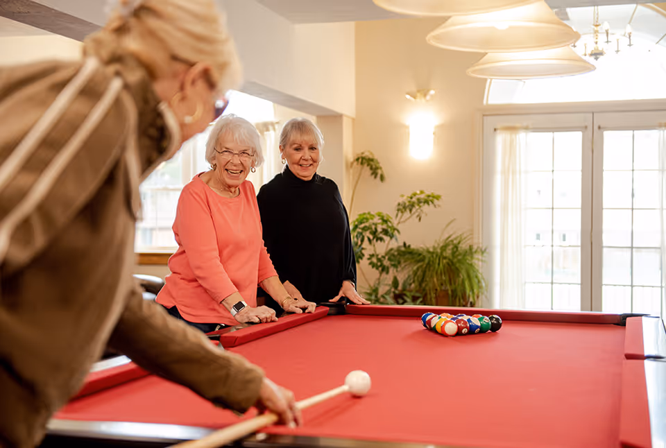 Three elderly women playing pool on a red felt pool table in a bright room with large windows and plants. Two women are standing at the side of the table smiling, while the third woman is taking a shot with a pool cue.