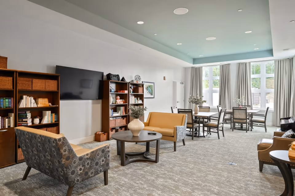 A spacious and well-lit living room area with large windows covered by light curtains. The room features a patterned armchair, a yellow loveseat, a round coffee table with a vase, bookshelves filled with books and decorative items, a wall-mounted flat-screen TV, and several tables with chairs arranged for seating or activities.