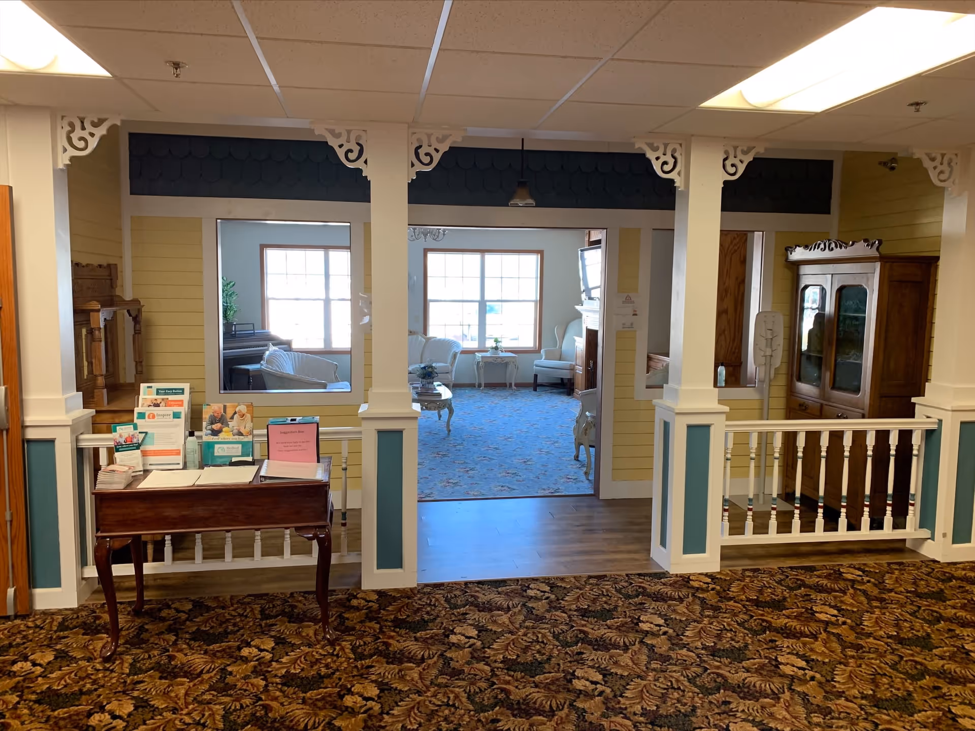 Interior view of a senior living facility showing a small wooden table with brochures and a suggestion box in the foreground, decorative white pillars and railings, and a cozy sitting area with armchairs, a piano, and large windows in the background.