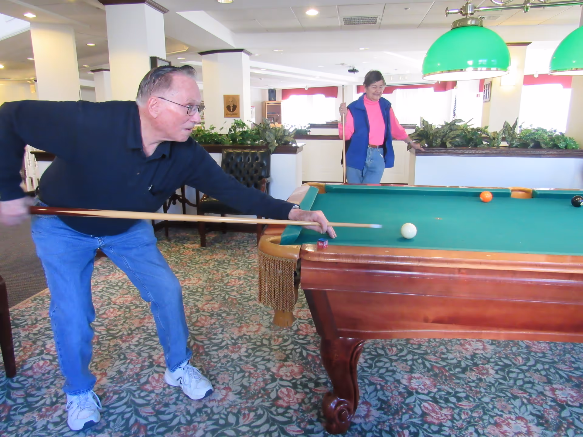 Two elderly individuals playing pool in a recreational room with floral carpet and green hanging lights above the pool table. One person is taking a shot while the other watches and smiles.