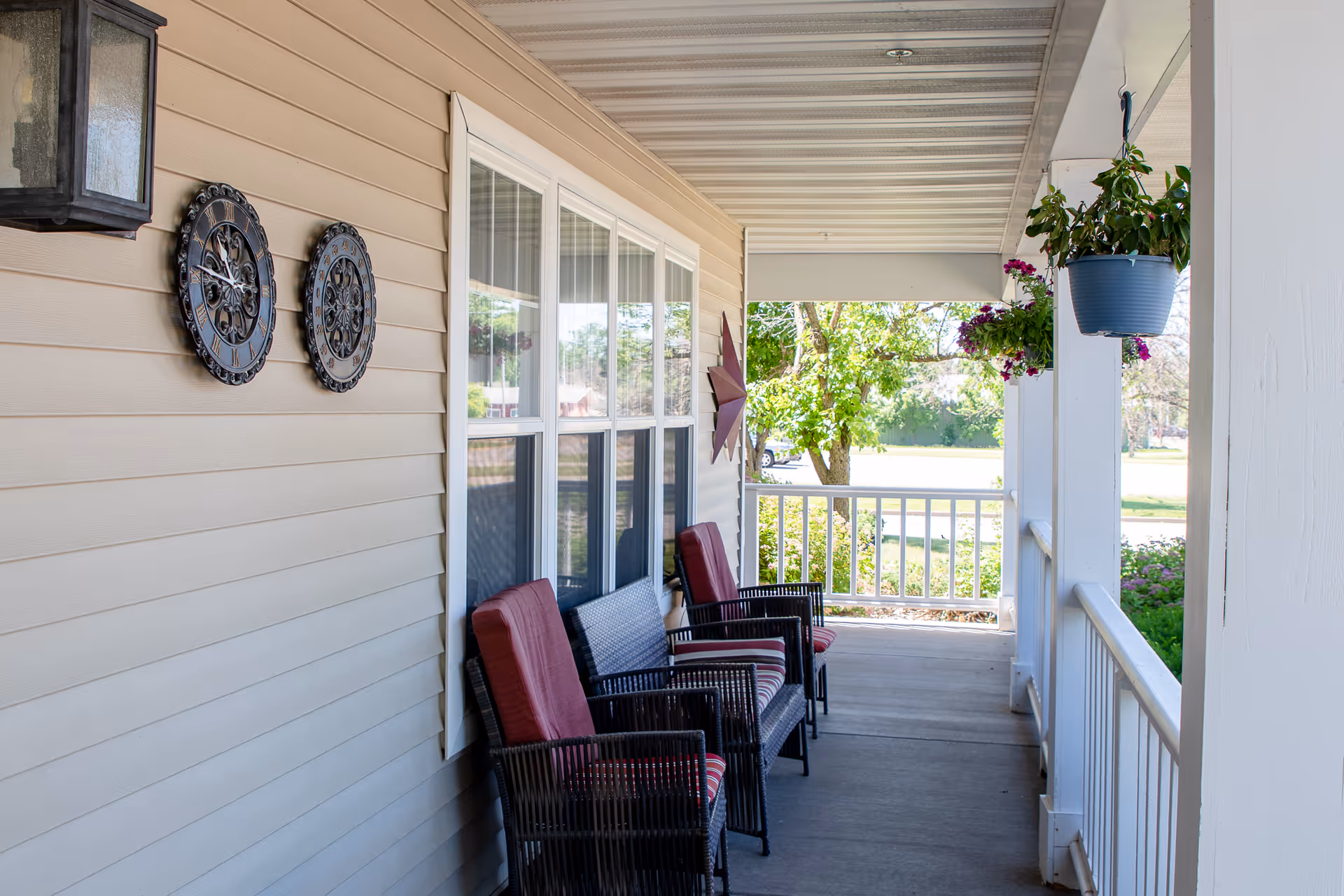 A covered front porch with wicker chairs topped with red cushions, hanging plants, decorative wall pieces, and a white railing.