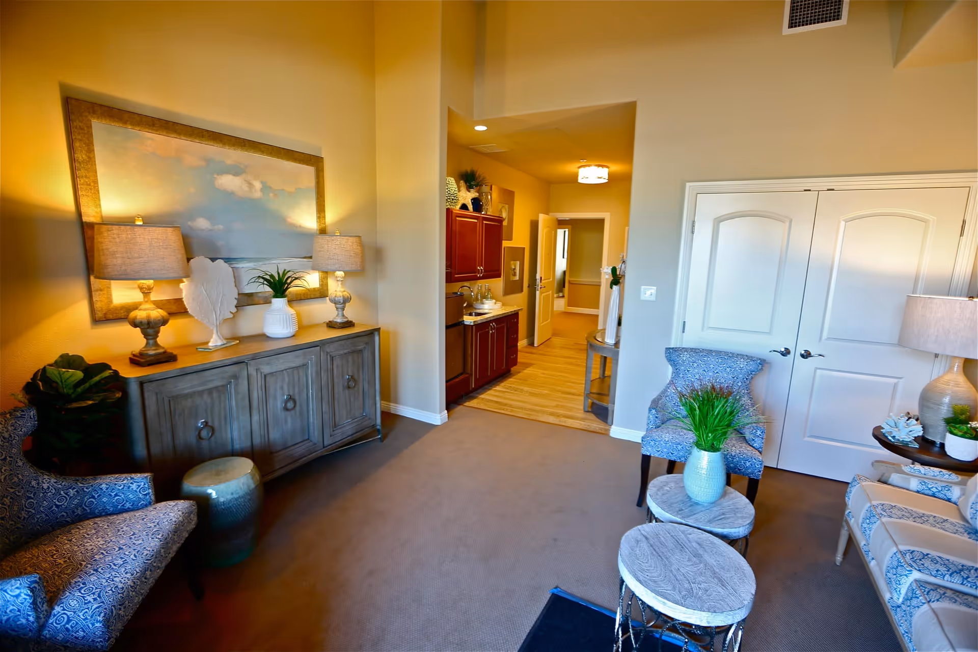 A cozy living room area with patterned blue armchairs, a beige sofa with blue accents, and a set of three round nesting tables with a green plant in a white vase on top. There is a wooden sideboard with two lamps, decorative items, and a large framed painting of a cloudy sky above it. The room has beige walls, carpeted flooring, and a doorway leading to a hallway with wooden flooring and cabinetry.