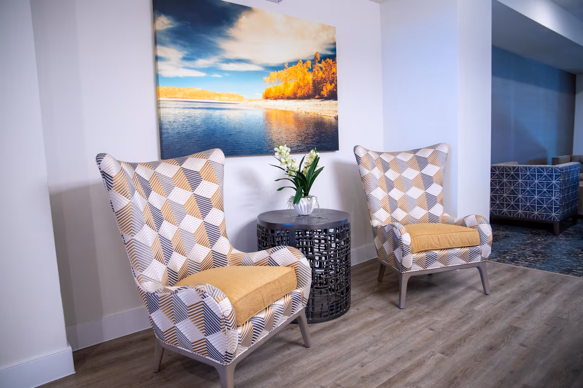 Two patterned armchairs with yellow cushions are positioned on either side of a small round black metal table with a glass vase holding white flowers. Behind the chairs is a wall with a large framed photograph of a lake and autumn trees under a partly cloudy sky. The floor is wood, and part of a blue patterned sofa is visible in the background.