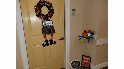 A decorated apartment door in a hallway with a Halloween wreath showing striped witch legs and small seasonal signs nearby.