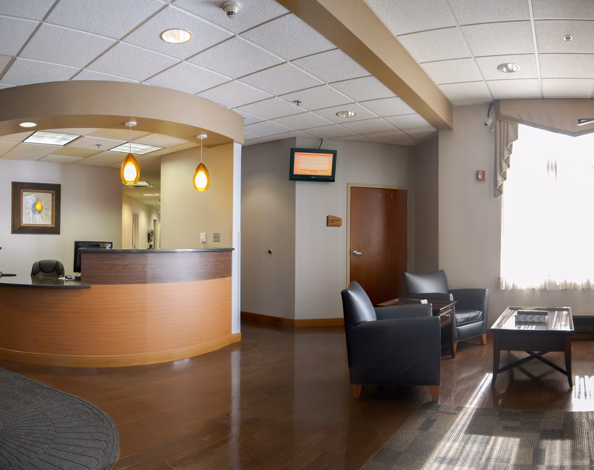 Reception area with a curved wooden desk, two hanging pendant lights, a black office chair behind the desk, and a hallway leading further inside. To the right, there are two black armchairs and a wooden coffee table in front of a large window with sheer curtains letting in natural light.