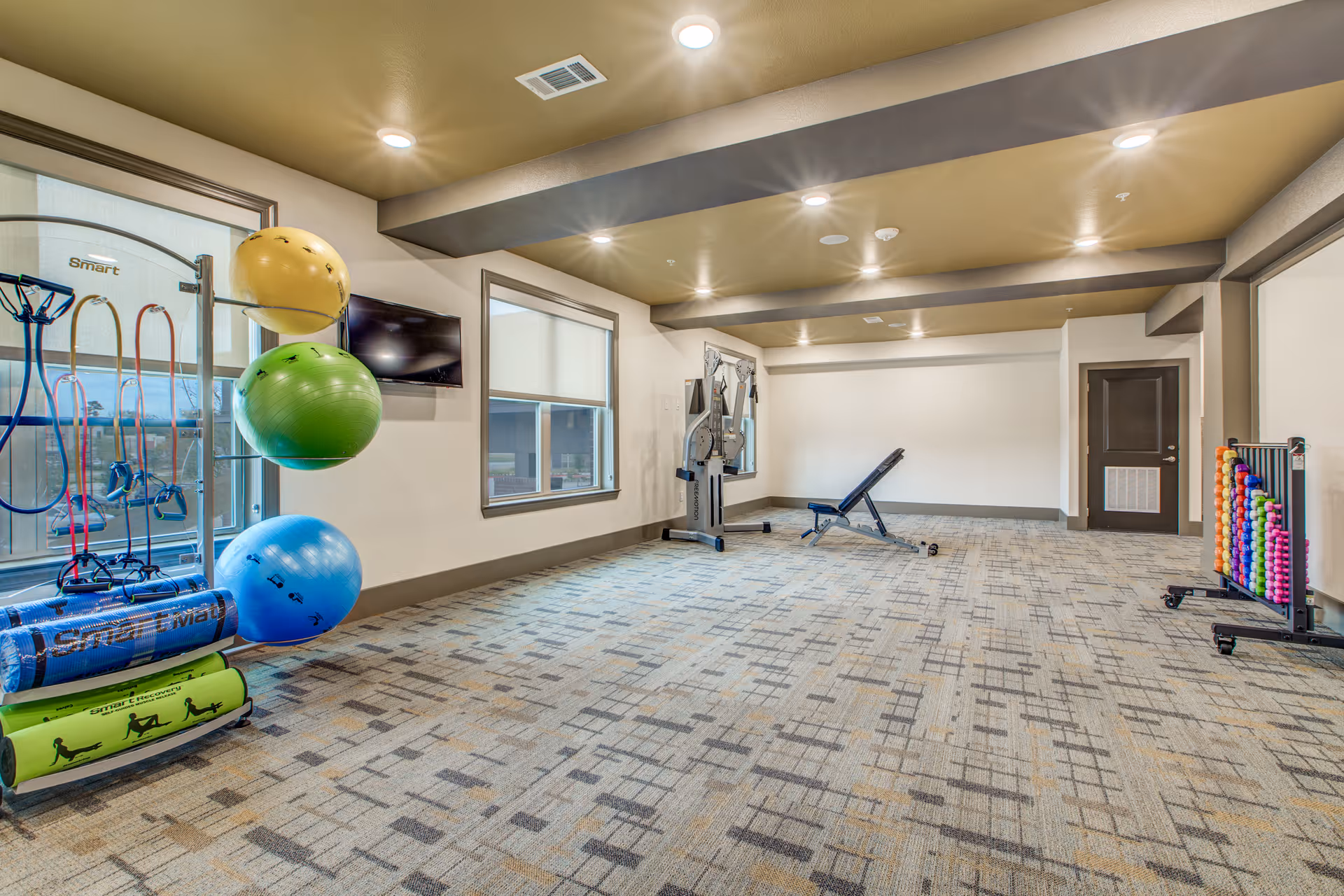A spacious fitness room with patterned carpet flooring, beige walls, and recessed lighting. The room contains various exercise equipment including colorful exercise balls, resistance bands, a weight bench, a multi-function workout machine, and racks holding small dumbbells. There are two windows with blinds and a wall-mounted TV.