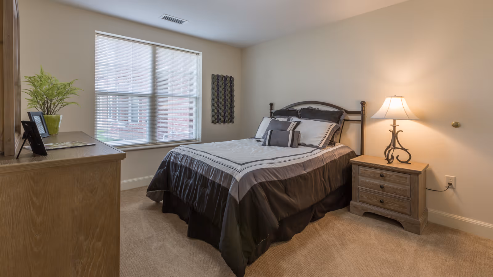 A cozy bedroom with a neatly made bed featuring black and white bedding, a wooden nightstand with a decorative lamp, a wooden dresser with a potted plant and picture frames, and a window with blinds letting in natural light.