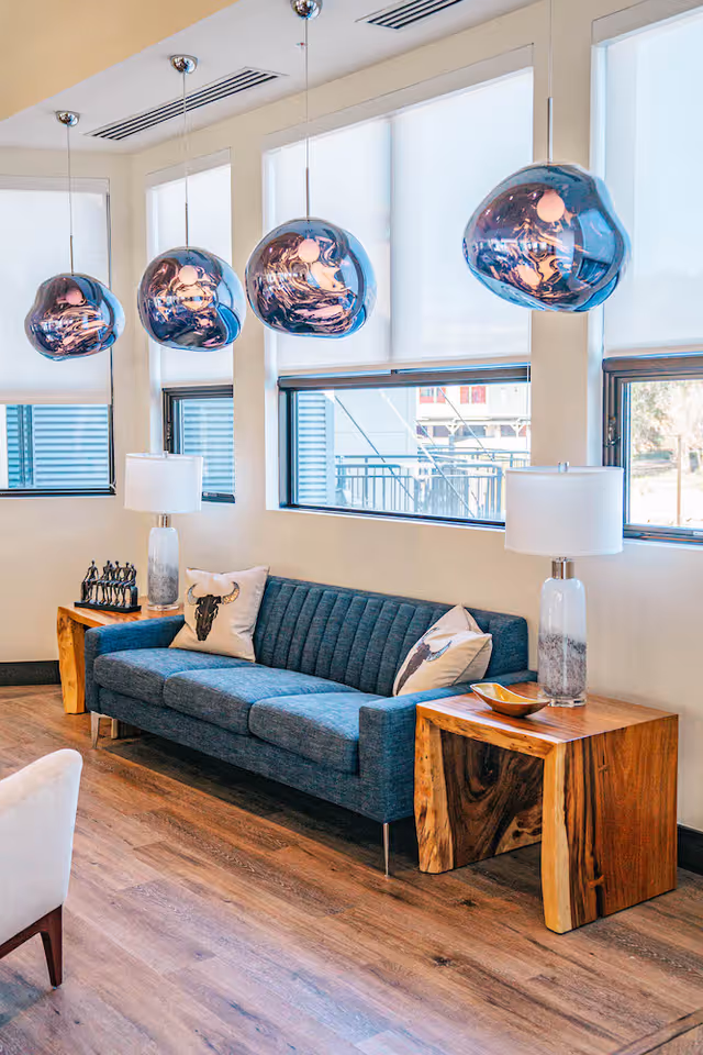 A modern living room area with a blue upholstered sofa adorned with two decorative pillows featuring animal designs. On each side of the sofa are wooden side tables with natural wood grain patterns, each holding a glass table lamp with a white shade. Above the sofa hang four unique, reflective pendant lights with a blue tint. The room has large windows with white roller shades, and the floor is covered with wood-style flooring.