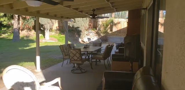 Covered outdoor patio area with a ceiling fan, a glass-top table surrounded by cushioned chairs, and additional seating including armchairs. The patio overlooks a grassy yard with trees and a wooden fence in the background.