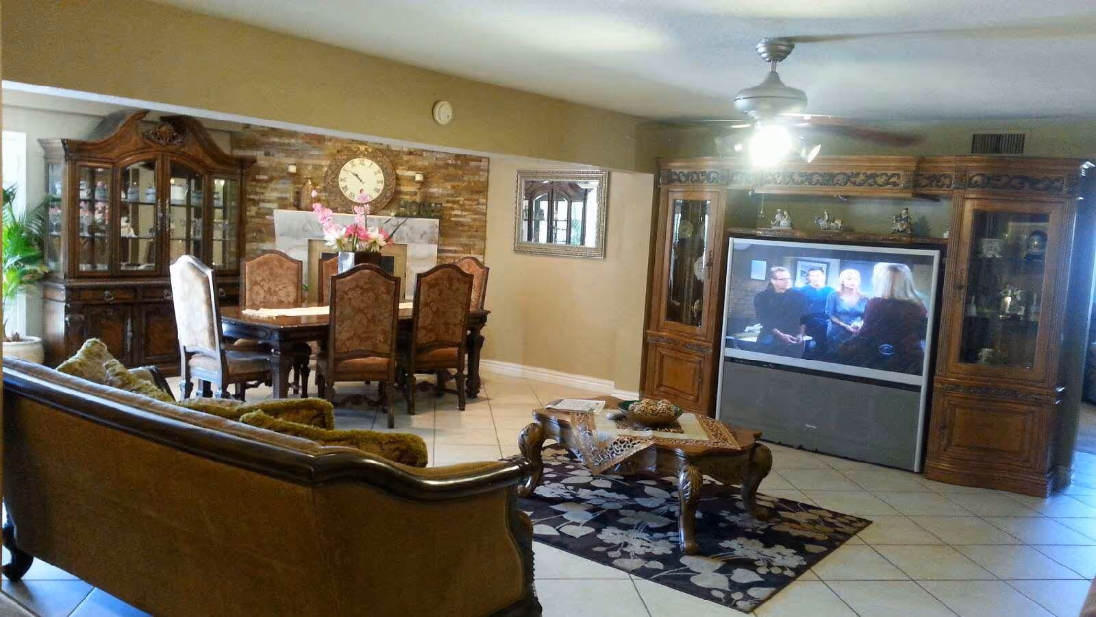 A combined living and dining room area with classic wooden furniture. The living room features a large wooden entertainment center with a television, a coffee table with decorative items, and a sofa with green cushions. The dining area has a wooden dining table with six upholstered chairs, a china cabinet, a wall clock above a fireplace, and a large mirror on the wall. The floor is tiled, and there is a ceiling fan with lights.