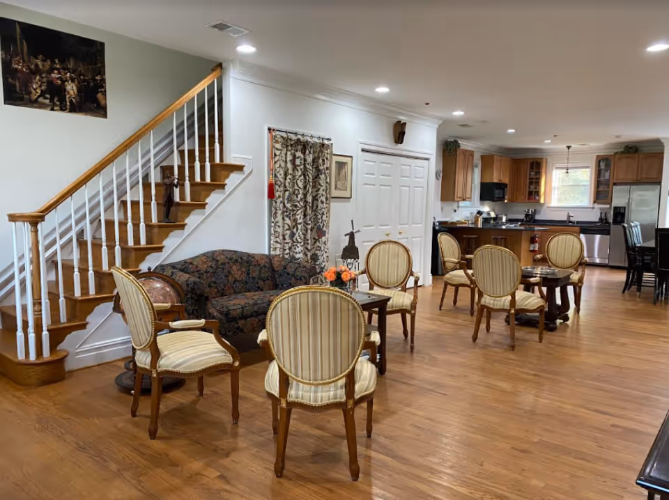 A spacious common area in an assisted living facility featuring a wooden staircase with white spindles, a floral patterned sofa, several striped upholstered chairs arranged around small tables, and a kitchen area with wooden cabinets, stainless steel appliances, and a window letting in natural light.