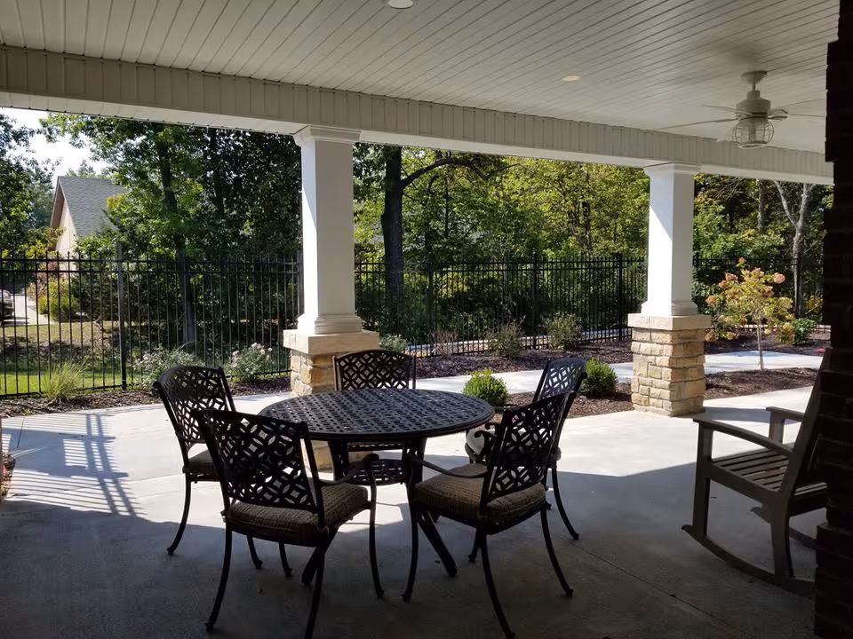 Covered outdoor patio area with a round metal table and four cushioned chairs, surrounded by white columns with stone bases. In the background, there is a black metal fence, trees, and greenery under a sunny sky.