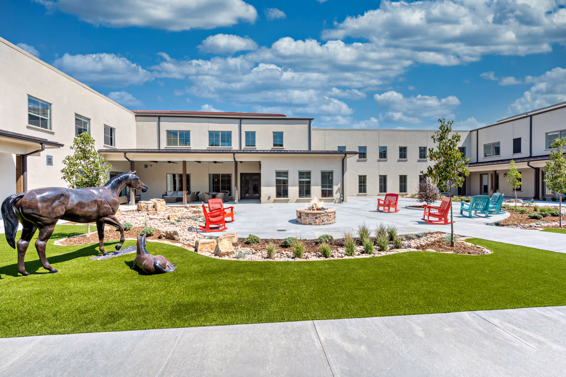 Outdoor courtyard area of StoneCreek of Flying Horse Senior Living featuring a green lawn with bronze horse statues, a circular fire pit surrounded by red and teal chairs, small trees, and a two-story beige building under a partly cloudy blue sky.