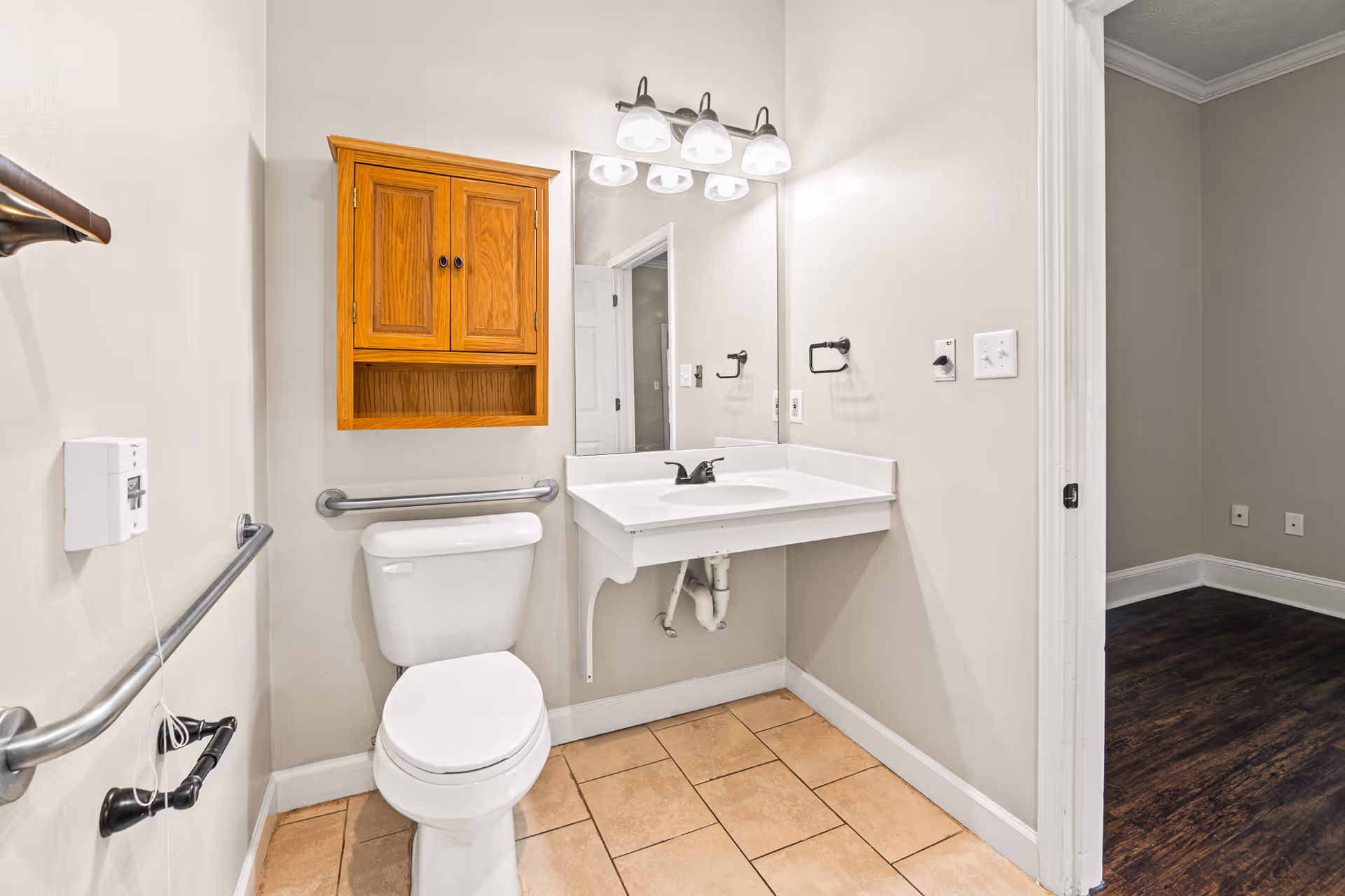 A clean bathroom with a white toilet, a wall-mounted wooden cabinet above the toilet, a white sink with a large mirror and three-light fixture above it. The bathroom has beige tiled flooring and beige walls with metal grab bars installed near the toilet for accessibility. The bathroom opens into a room with dark wood flooring.