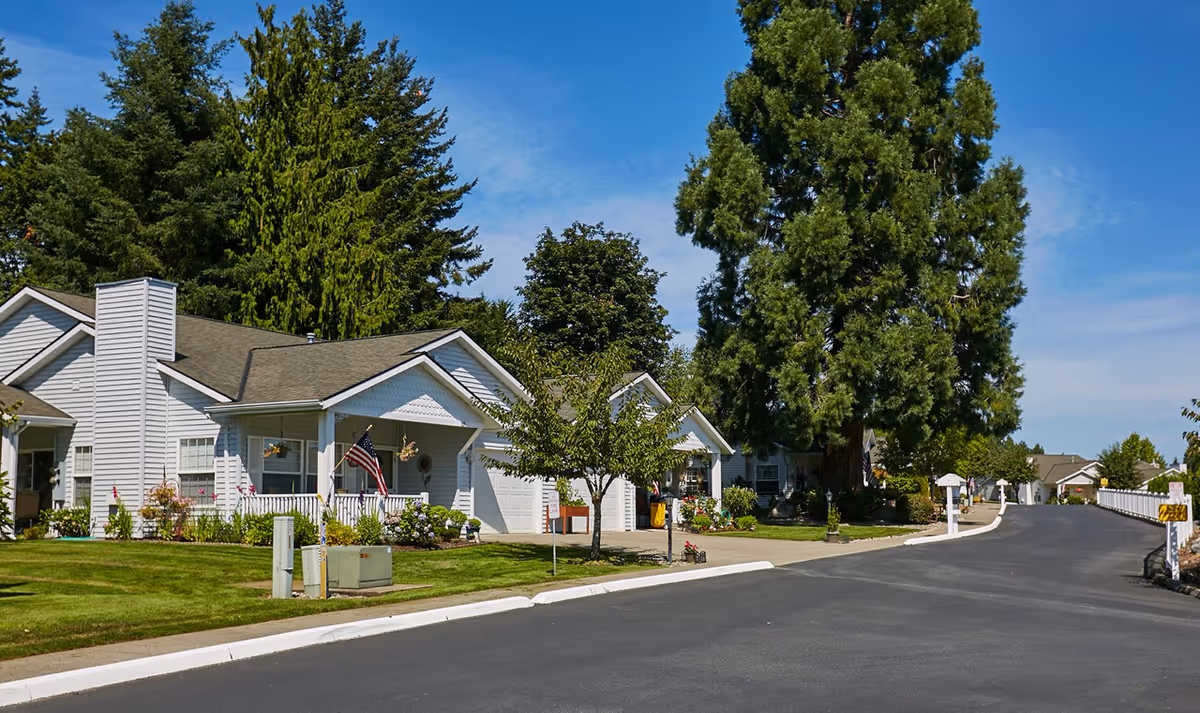 A paved road curves through a senior living community with single-story white houses, well-maintained lawns, trees, and clear blue sky.