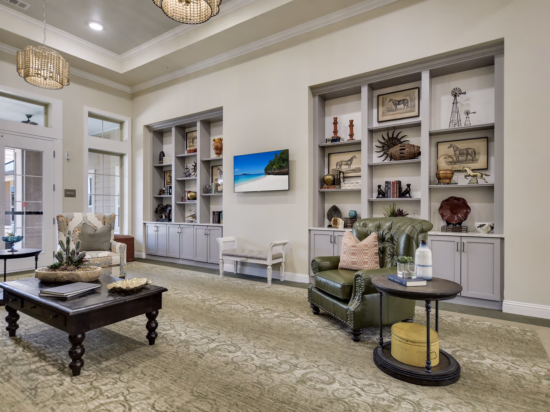A cozy living room area in a senior living facility with a green leather armchair, a patterned armchair, a wooden coffee table, and built-in shelves displaying decorative items and framed pictures of horses. The room has a large window and glass door letting in natural light, with a beige carpet and neutral wall colors.