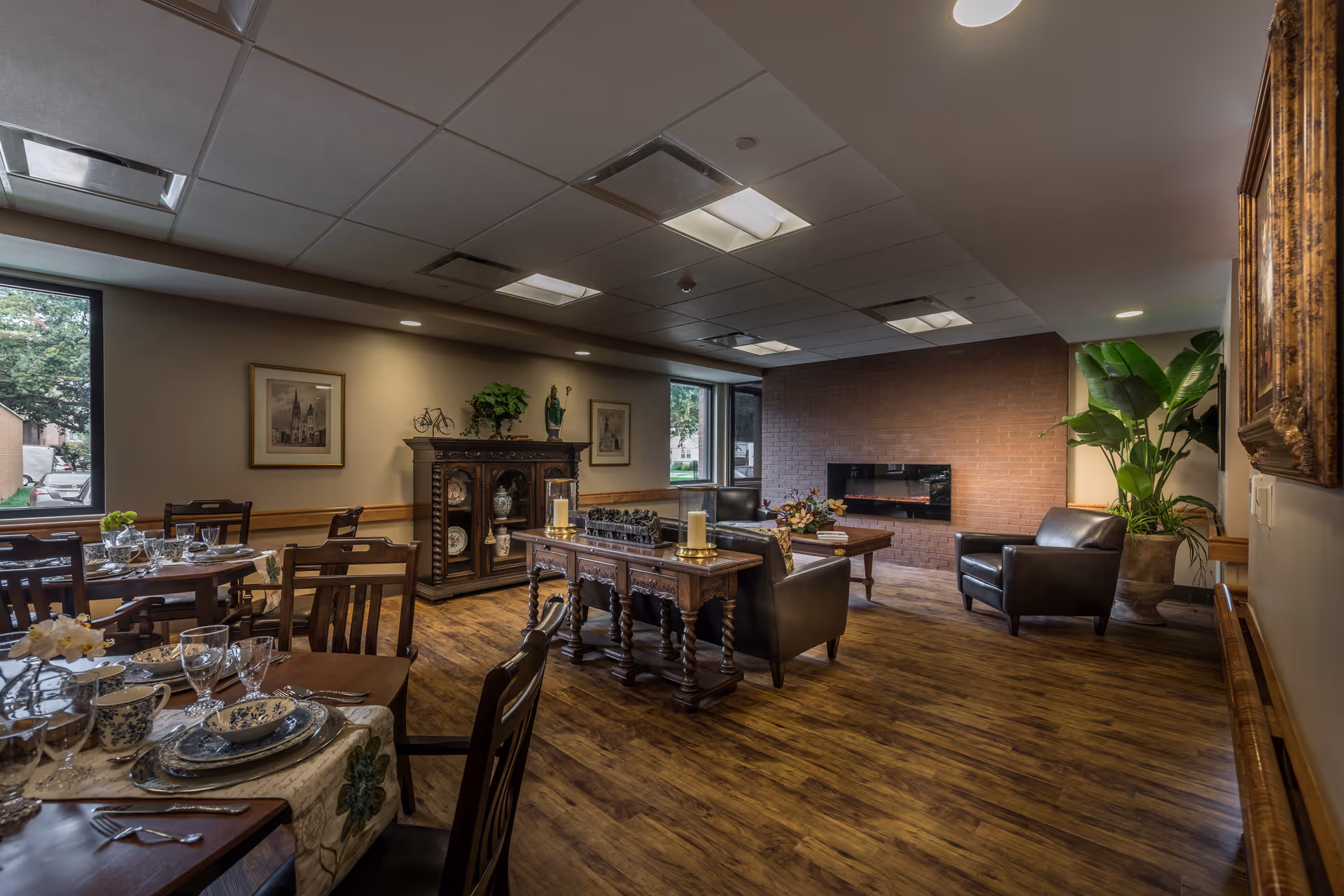Warmly lit senior living common room with dining tables set for a meal, a seating area with leather chairs and a fireplace, and decorative furnishings.