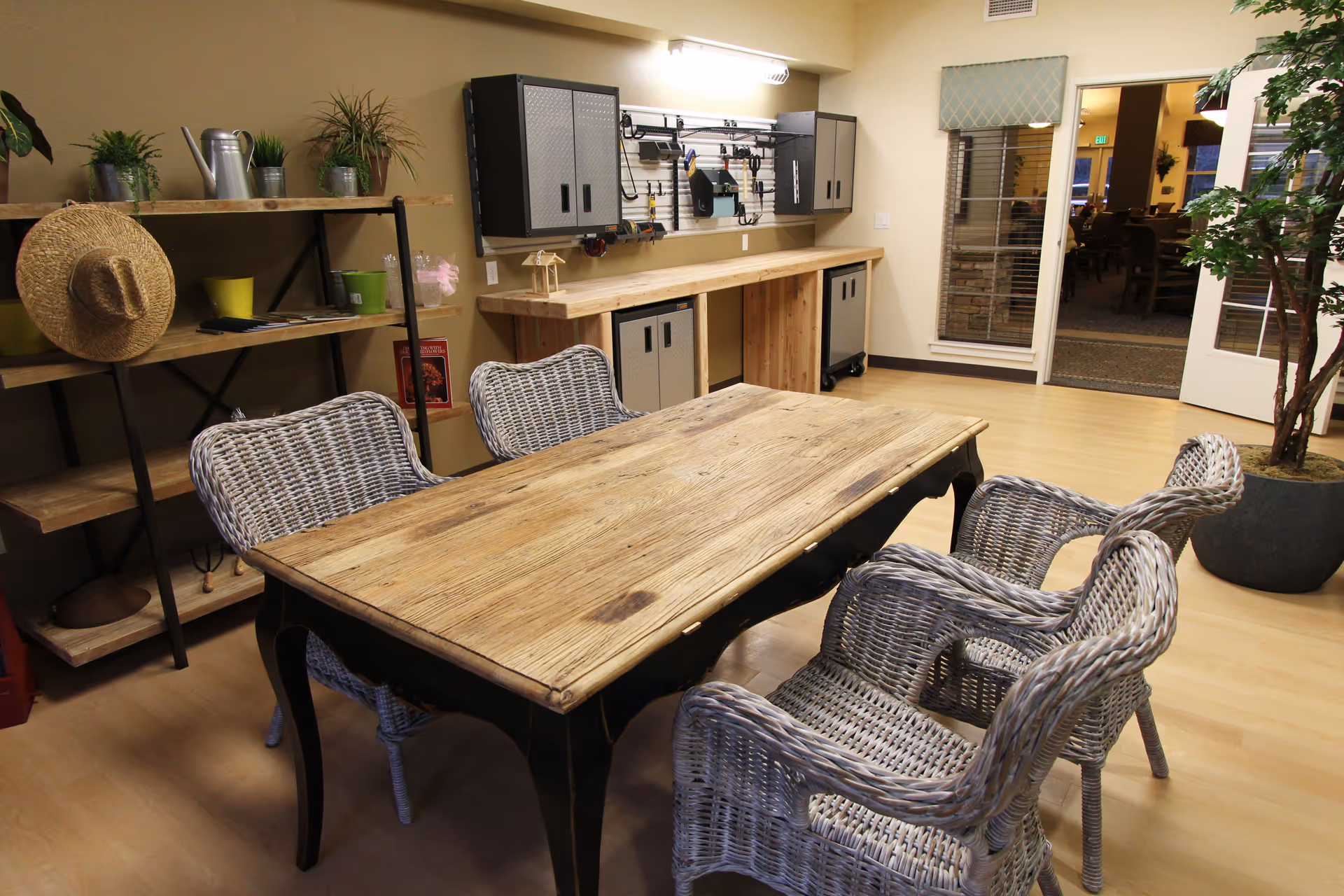 A cozy interior room featuring a wooden table surrounded by four wicker chairs. To the left, there is a shelving unit with plants, a straw hat, and various items. On the far wall, there is a workbench with cabinets and tools hanging above it. The room has light wood flooring and a large potted plant near a glass door leading to another room with dining tables and chairs.