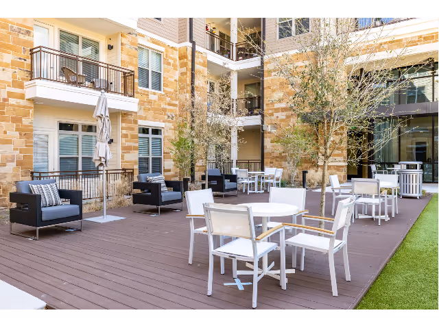 Outdoor patio area at a senior living facility with white round tables and chairs, black cushioned lounge chairs, small trees, and a building with balconies and large windows in the background.