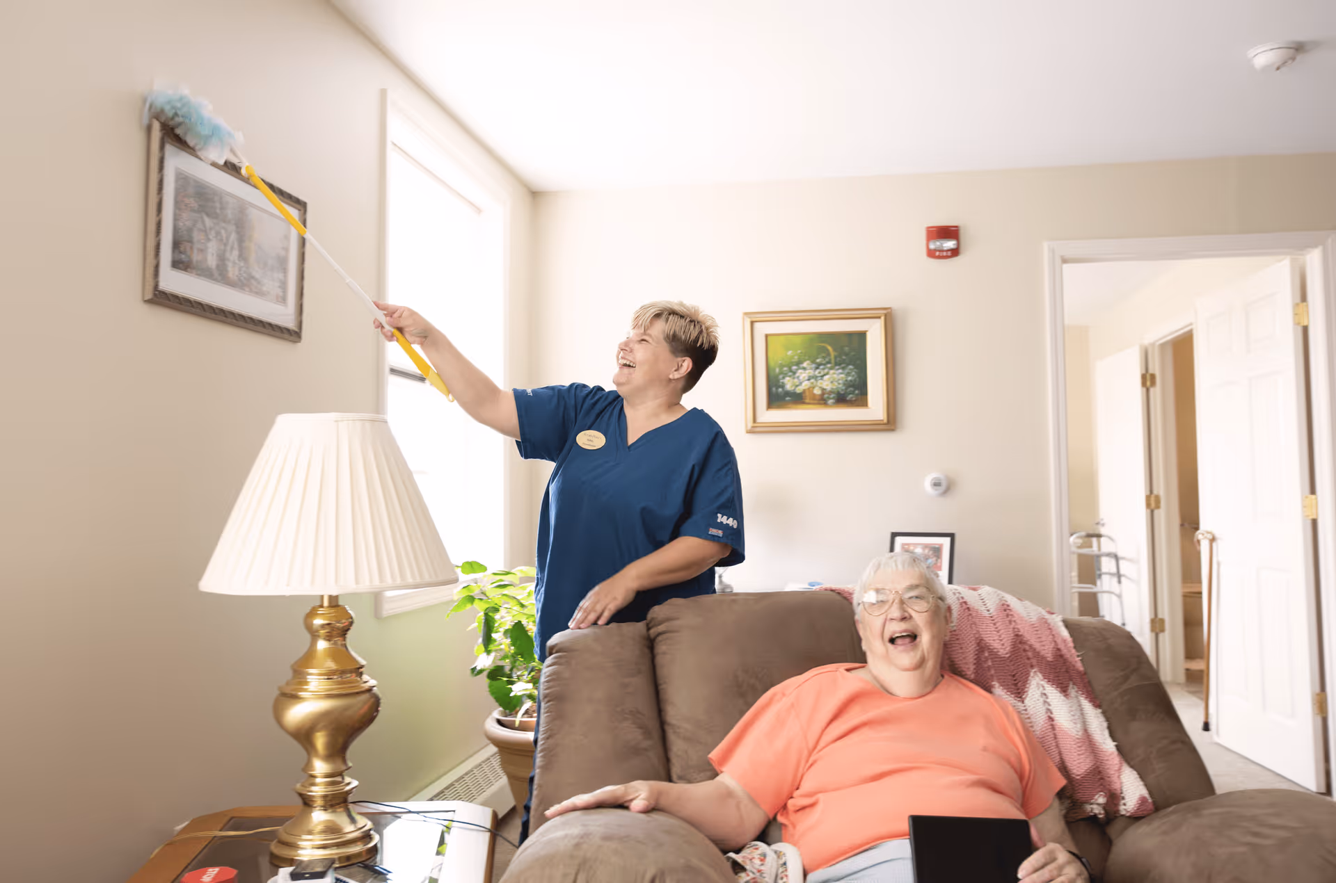 A caregiver in blue scrubs dusts a picture frame on the wall with a duster while an elderly woman with glasses and a peach shirt sits on a brown recliner chair smiling. The room has beige walls, a window with natural light, a brass table lamp, a potted plant, and framed artwork on the walls.