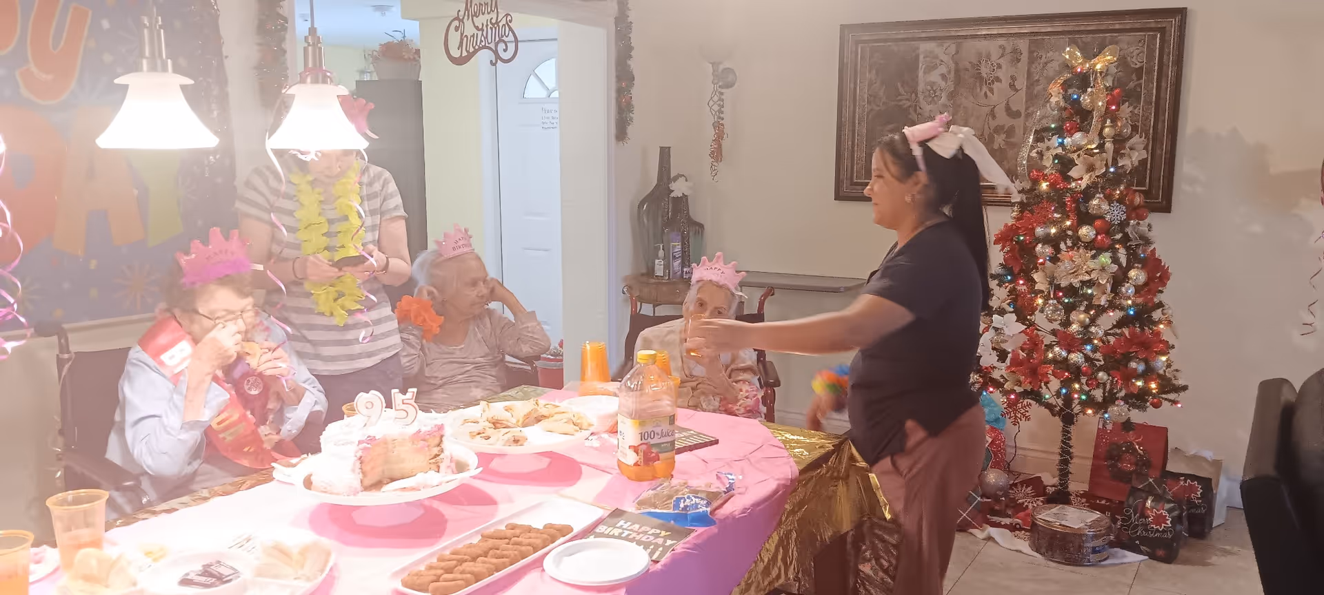 A group of elderly women wearing pink birthday crowns sitting around a table with birthday cake and snacks. A woman stands near the table, interacting with them. The room is decorated with a Christmas tree and holiday decorations, including a 'Merry Christmas' sign and a birthday banner.