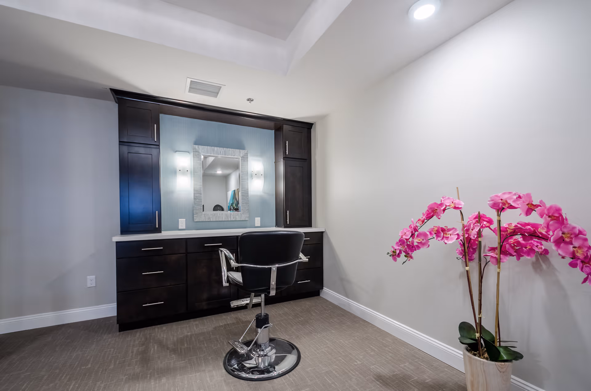 A modern vanity area with dark wood cabinets, a large mirror with two wall sconces on either side, and a black salon chair in front. To the right, there is a potted plant with vibrant pink flowers against a plain white wall.
