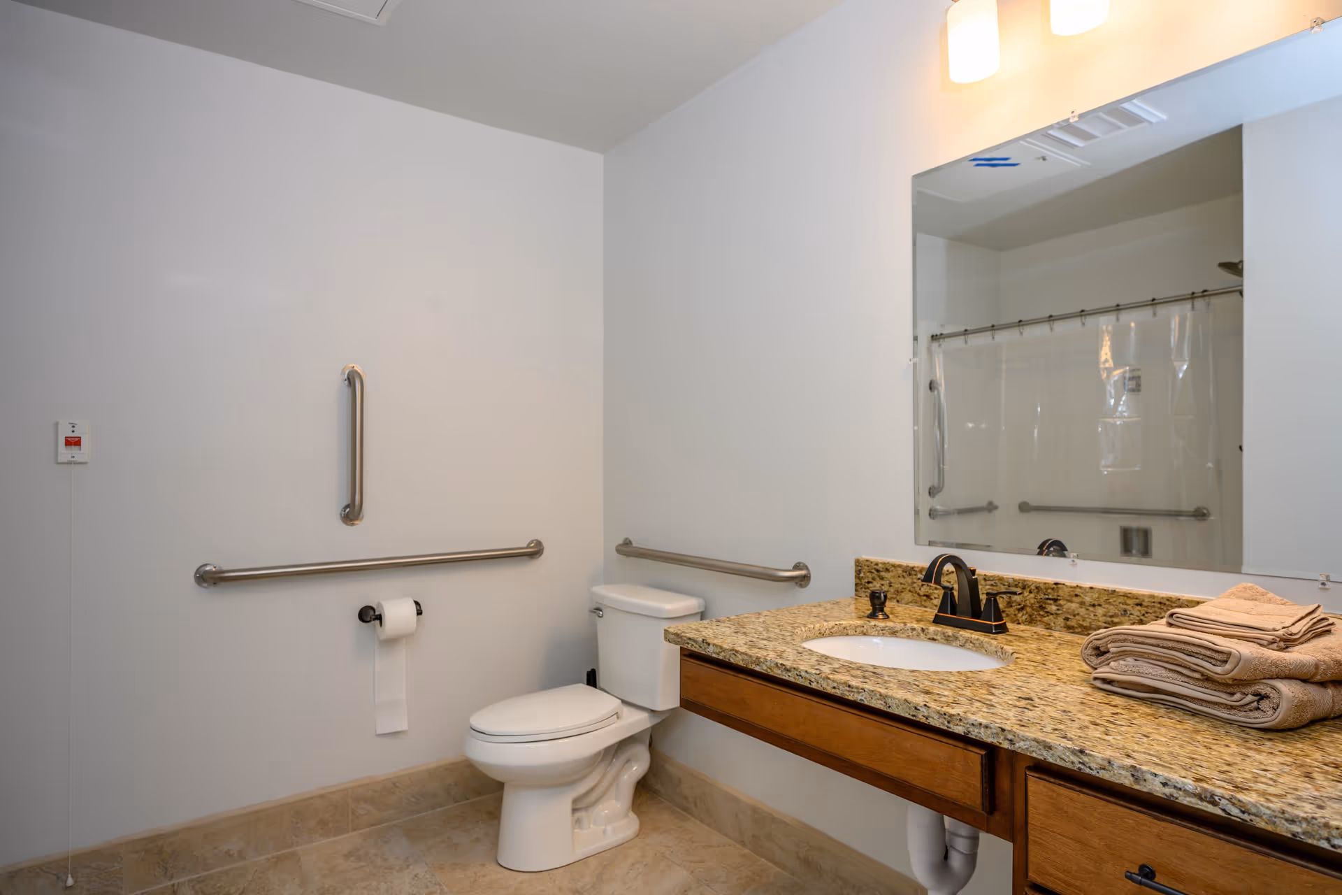 A clean and accessible bathroom with a toilet, granite countertop with sink, folded towels, a large mirror, and grab bars on the walls. The shower area is visible in the mirror reflection with a clear shower curtain and additional grab bars.