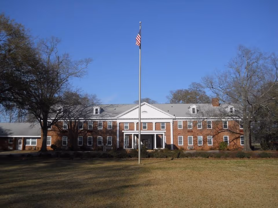 Front exterior view of a large two-story brick building with white trim and a central entrance with columns. An American flag flies on a flagpole in front of the building. Leafless trees flank the building under a clear blue sky.