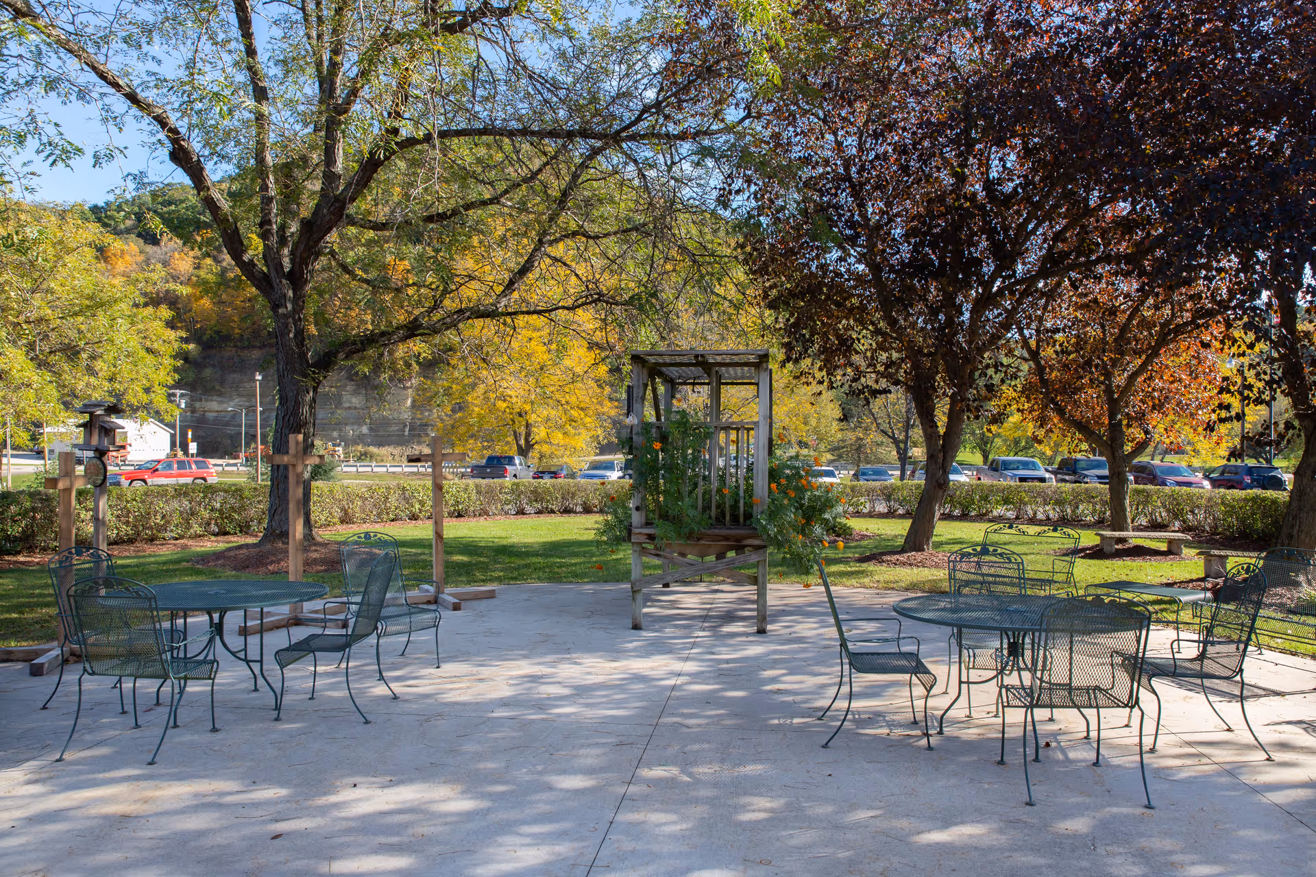 Outdoor patio area with metal tables and chairs on a concrete surface, surrounded by trees with green and autumn-colored leaves. A wooden planter structure with flowers is in the center background, and parked cars and a rocky hill are visible beyond the hedge bordering the patio.